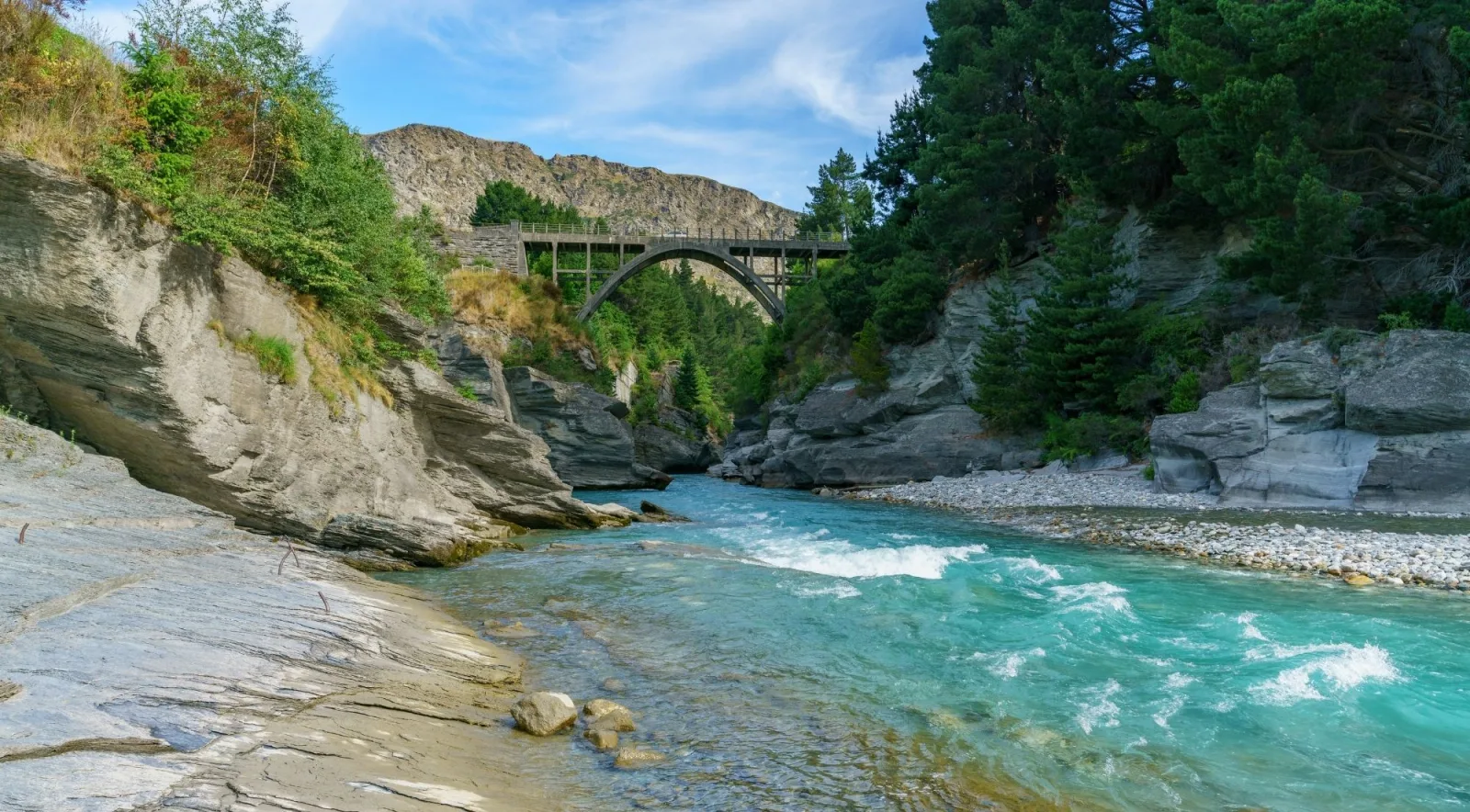 A turquoise river running between steep canyon walls with a bridge far overhead
